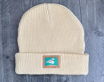 Peut inclure: Un bonnet en tricot couleur crème avec un revers plié. Au centre du revers se trouve un écusson rectangulaire sur fond sarcelle représentant deux petits oiseaux blancs et gris sur une surface de sable beige.