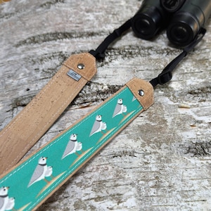 May include: A teal and tan camera strap with a bird pattern. The strap has a cork section and a teal section with white birds and orange accents. The strap is connected to a pair of black binoculars.
