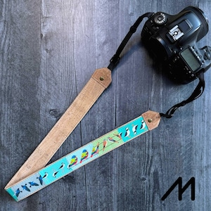 Puede incluir: Una correa de cámara con diseño de corcho y tela. La parte de corcho es marrón claro, mientras que la tela presenta un fondo turquesa con pájaros coloridos. Una cámara negra está sujeta a la correa.