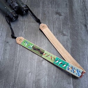 Puede incluir: Una correa para binoculares con temática de aves con un diseño de corcho y tela. La correa presenta un fondo verde con varias ilustraciones de aves en diferentes colores. La correa está conectada a un par de binoculares negros.