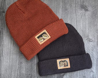 Peut inclure: Deux bonnets en tricot à revers, l'un orange rouille et l'autre gris anthracite. Chaque bonnet comporte un écusson rectangulaire sur le revers. Le bonnet orange a un écusson avec deux hiboux, tandis que le gris a un groupe de hiboux.