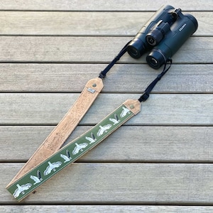 May include: A brown leather camera strap with a green fabric insert featuring a repeating pattern of white birds. The strap is attached to a pair of black binoculars with the word "Vortex" printed on the side.