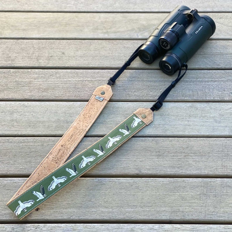 May include: A brown leather camera strap with a green fabric insert featuring a repeating pattern of white birds. The strap is attached to a pair of black binoculars with the word "Vortex" printed on the side.