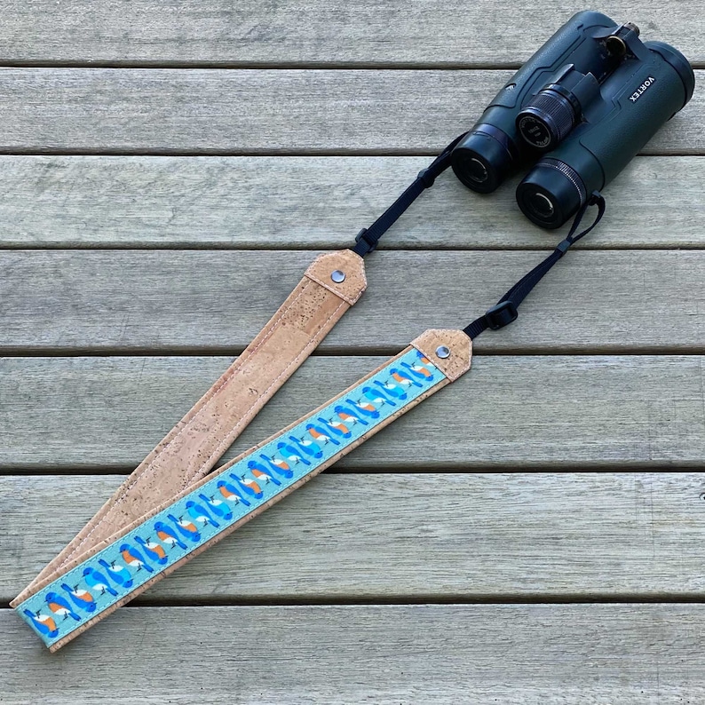 May include: A brown cork and blue patterned camera strap with black adjustable straps. The strap is laying on a wooden surface.