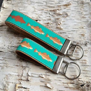 May include: Two keychains with a teal fabric featuring orange fish. The keychains have a brown leather backing and silver-colored metal hardware. The keychains are on a light-colored, textured surface.