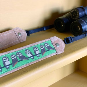 May include: A camera strap made of cork and fabric with a green background and a repeating pattern of owls. The strap is attached to a pair of binoculars.