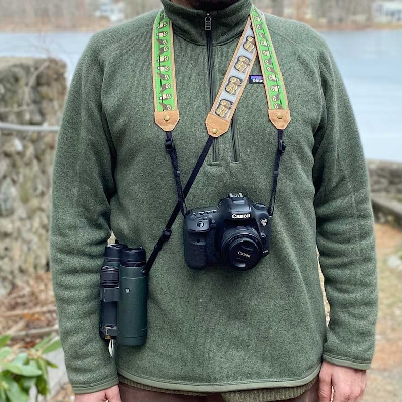 May include: A Canon EOS camera with a lens is suspended by a patterned camera strap. The strap features a repeating design of brown owls on a green background. A pair of binoculars hangs on the left side. The person is wearing a green fleece jacket.