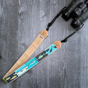 Peut inclure: Une paire de jumelles repose sur une surface en bois grise, à côté de deux sangles décoratives. L'une est en liège brun clair, l'autre présente un motif bleu et noir avec des illustrations de poissons. Les sangles sont conçues pour tenir les jumelles.