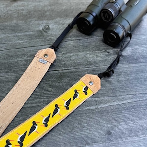 Pode incluir: Close-up de binóculos com uma alça amarela com um padrão de pássaros pretos e uma alça de cortiça. A alça amarela tem um padrão repetido de pássaros pretos com detalhes brancos e vermelhos. A alça de cortiça tem uma pequena etiqueta de metal.