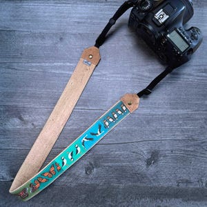 Puede incluir: Una correa de cámara con un diseño de corcho y tela. La parte de corcho es de color marrón claro, mientras que la tela presenta un fondo turquesa con ilustraciones de mariposas y pájaros. Una cámara negra está sujeta a la correa.