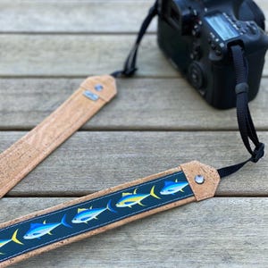 May include: A camera strap with a blue and white fish pattern on a dark blue background. The strap is made of cork and has a black strap attached to a black camera.