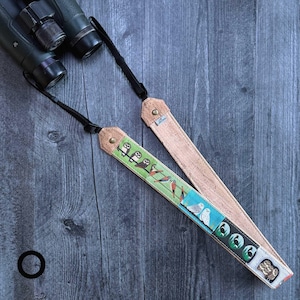 May include: Two birding binocular straps. One strap features a green background with bird illustrations, while the other is a natural cork color. Both straps have black cords and are attached to a pair of binoculars.