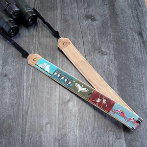 May include: A decorative bird-themed binocular strap with a cork backing and black adjustable straps. The strap features colorful illustrations of owls, birds on a wire, and other bird species. The strap is designed to be attached to binoculars.