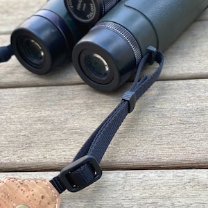 May include: Close-up of a pair of binoculars with a black strap. The binoculars have a green and black body with the text "TALON" visible. The strap is attached to a cork and leather piece.