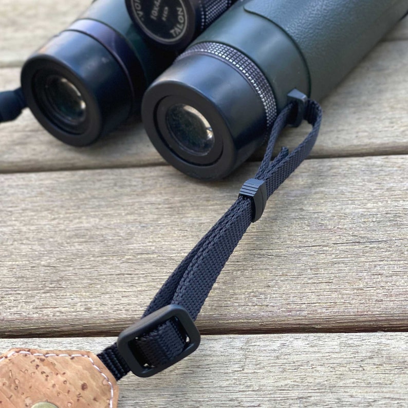 May include: Close-up of a pair of binoculars with a black strap. The binoculars have a green and black body with the text "TALON" visible. The strap is attached to a cork and leather piece.