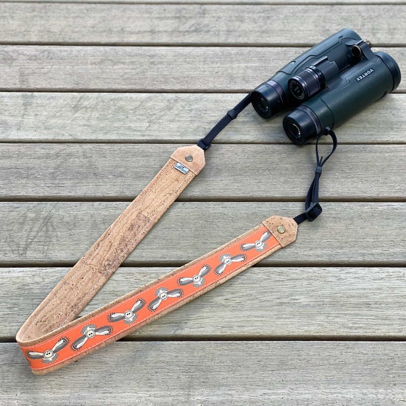 May include: A pair of green and black binoculars with a cork strap. The strap has an orange section with a propeller design. The binoculars are resting on a wooden surface.