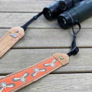 May include: A cork and orange owl-themed binocular strap. The strap features a repeating pattern of white owls on an orange background, with cork ends and black adjustable straps. Binoculars are in the background.