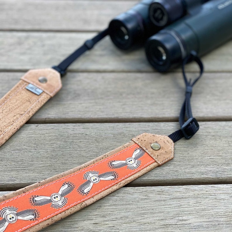 May include: A cork and orange owl-themed binocular strap. The strap features a repeating pattern of white owls on an orange background, with cork ends and black adjustable straps. Binoculars are in the background.