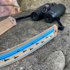 May include: A brown leather camera strap with a blue and white pattern of black birds. The strap is attached to a pair of binoculars.