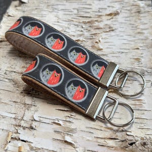 May include: Two keychains with a repeating pattern of cat faces in circles on a dark gray background. The keychains have a cork-like material on the edges and silver-colored metal hardware with key rings.