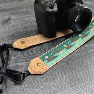 Puede incluir: Una cámara DSLR negra con una correa para cámara con temática de patos. La correa presenta un fondo verde con un patrón de patos nadando en un estanque. La correa está hecha de corcho y tela.