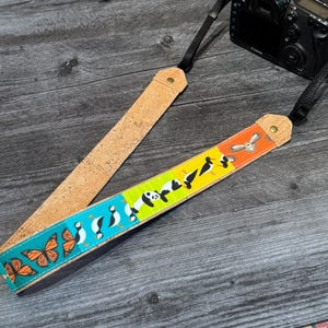 May include: A camera strap with a cork shoulder pad and a colorful patterned section. The patterned section features butterflies, penguins, pandas, and birds. The strap is attached to a black Canon camera.