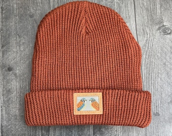 Peut inclure: Un bonnet en tricot couleur rouille avec un revers plié. Au centre du revers se trouve un écusson rectangulaire illustrant deux petits oiseaux au plumage orange, bleu et blanc, se faisant face sur un fond clair.
