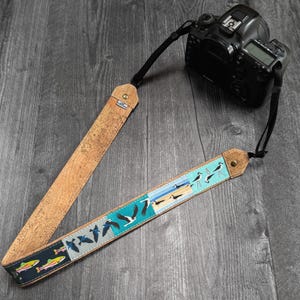 Może przedstawiać: Pasek do aparatu z korka i tkaniny. Część korkowa jest jasnobrązowa, a tkanina ma niebieski i turkusowy wzór z ilustracjami ptaków i ryb. Czarny aparat jest przymocowany do paska.