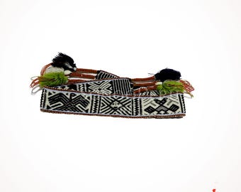 May include: A woven belt with a geometric pattern in black and white, accented with brown trim and tassels in black and green. The belt features intricate designs and is likely handmade.