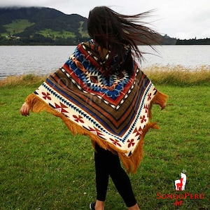 May include: A patterned poncho in brown, blue, red, and white, with a fringed edge. The poncho features geometric designs and is worn outside. The background shows a lake and green grass.