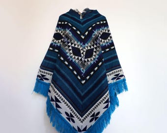 May include: A blue poncho featuring a geometric pattern in shades of navy, light blue, white, and grey. The garment has a V-shaped neckline with a single button closure at the top and is finished with long blue fringe along the bottom hem.
