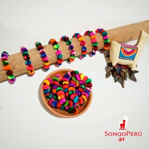 Puede incluir: Un montón de ribete de pompón hecho a mano de colores vivos, hecho de lana de alpaca, en una variedad de colores, incluyendo rojo, naranja, amarillo, verde, azul, morado y rosa. El ribete se muestra en una clavija de madera y en un pequeño cuenco de terracota. El ribete es perfecto para agregar un toque de color y textura a cualquier proyecto.