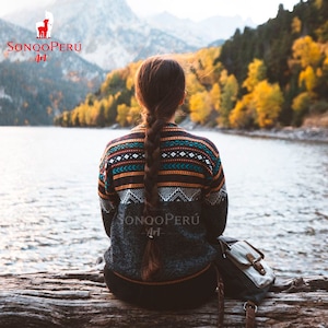 May include: A person wearing a grey sweater with a colourful geometric pattern sits on a log overlooking a lake. The sweater has the text "SONQO PERÚ Art" on the front. The person has long brown hair braided down their back.
