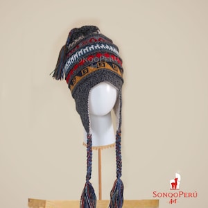 May include: Grey knitted hat with a colourful geometric pattern and long braided tassels. The hat has ear flaps and a pom-pom on top.