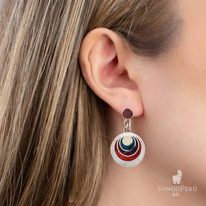 Könnte beinhalten: Ein Paar silberne Ohrringe mit einem runden Design. Die Ohrringe haben konzentrische Kreise aus rotem, weißem, blauem und grünem Emaille. Ein kleiner, dunkelroter Edelstein hängt an der Unterseite jedes Ohrrings.