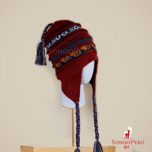 Può includere: Un cappello di lana rosso con un motivo geometrico colorato e lunghi pon pon intrecciati. Il cappello ha le alette per le orecchie e un pon pon in cima.