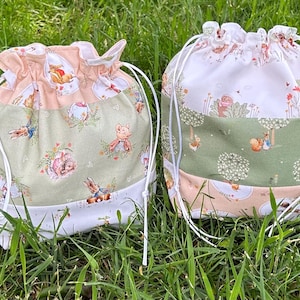 May include: Two drawstring bags with a whimsical, storybook design. One bag features a peach and green colour scheme with rabbit and floral patterns. The other bag has a white, green, and peach colour scheme with squirrel and tree patterns.