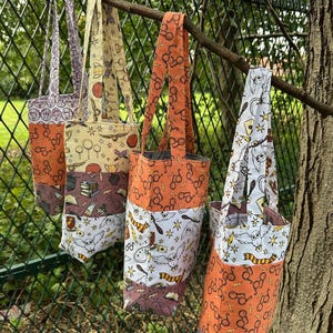 May include: Four tote bags hanging from a tree branch. The bags feature patterns of glasses, books, owls, and other themed designs. The bags have orange, white, and purple accents, with matching handles.
