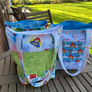 May include: Three Paddington Bear tote bags. The bags feature fabric panels with London-themed stamps and Paddington Bear illustrations in white, blue, and green. Each bag has a blue interior and white handles.