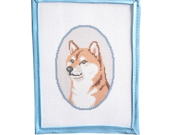 May include: A rectangular needlepoint canvas featuring a cross-stitched portrait of a Shiba Inu dog in shades of tan, white, and black. The dog is centered within an oval frame, and the entire canvas is edged with a light blue fabric binding.