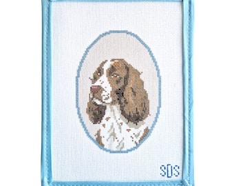 May include: A rectangular needlepoint canvas featuring a cross-stitched portrait of a brown and white spaniel dog inside a light blue oval border. The piece is finished with a light blue fabric binding and includes the initials SDS in the corner.