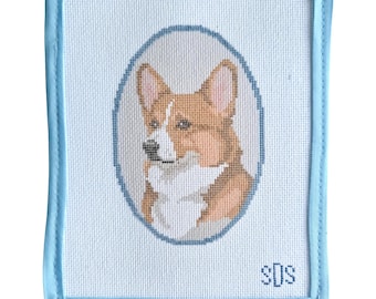 May include: A rectangular needlepoint canvas featuring a portrait of a brown and white Corgi dog inside a blue oval border. The canvas has a light blue fabric binding along the edges and the initials SDS stitched in blue in the bottom right corner.