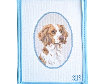 May include: A rectangular needlepoint canvas featuring a portrait of a brown and white dog inside a blue oval border. The canvas is finished with a light blue fabric binding on all four edges and includes the embroidered initials SDS in blue.