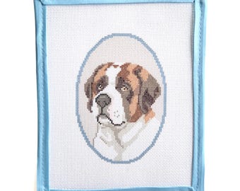 May include: A rectangular needlepoint canvas featuring a cross-stitched portrait of a Saint Bernard dog. The dog is centered within a light blue oval border, and the entire canvas is finished with a light blue fabric binding on all four edges.