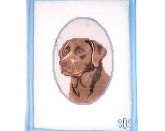 May include: A rectangular needlepoint canvas featuring a brown dog portrait centered within a light blue oval border. The canvas has a light blue fabric binding along the edges and the letters SOS stitched in the bottom right corner on white mesh.