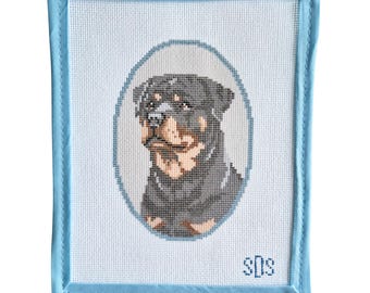 May include: A rectangular needlepoint canvas featuring a Rottweiler dog portrait centered within an oval frame. The dog has black and tan fur. The canvas has a light blue fabric border and the initials SDS stitched in blue in the bottom right corner.