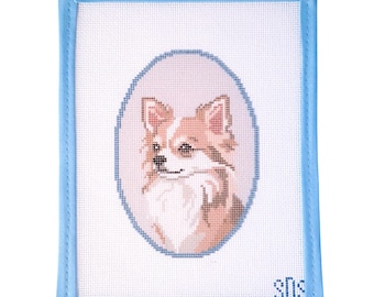 May include: A rectangular needlepoint canvas featuring a cross-stitched portrait of a long-haired dog with tan and white fur, centered within a blue oval border. The canvas is finished with a light blue fabric binding on all four edges.