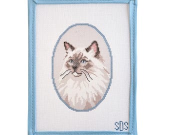 May include: A rectangular needlepoint canvas featuring a cross-stitched portrait of a long-haired cat with blue eyes inside an oval frame. The canvas is bordered by light blue fabric binding and includes the embroidered initials SDS in the corner.