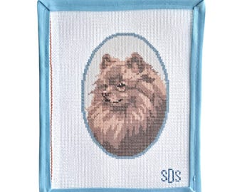 May include: A rectangular needlepoint canvas featuring a portrait of a brown Pomeranian dog inside a light blue oval frame. The canvas has a light blue fabric border and the letters SDS stitched in blue in the bottom right corner on white mesh.