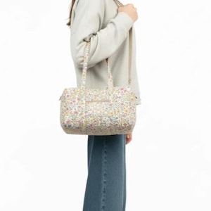 May include: A floral print handbag with a shoulder strap and top handles. The bag features a pattern of small flowers in shades of pink, yellow, and blue on a cream background. The bag is rectangular with a zipper closure.